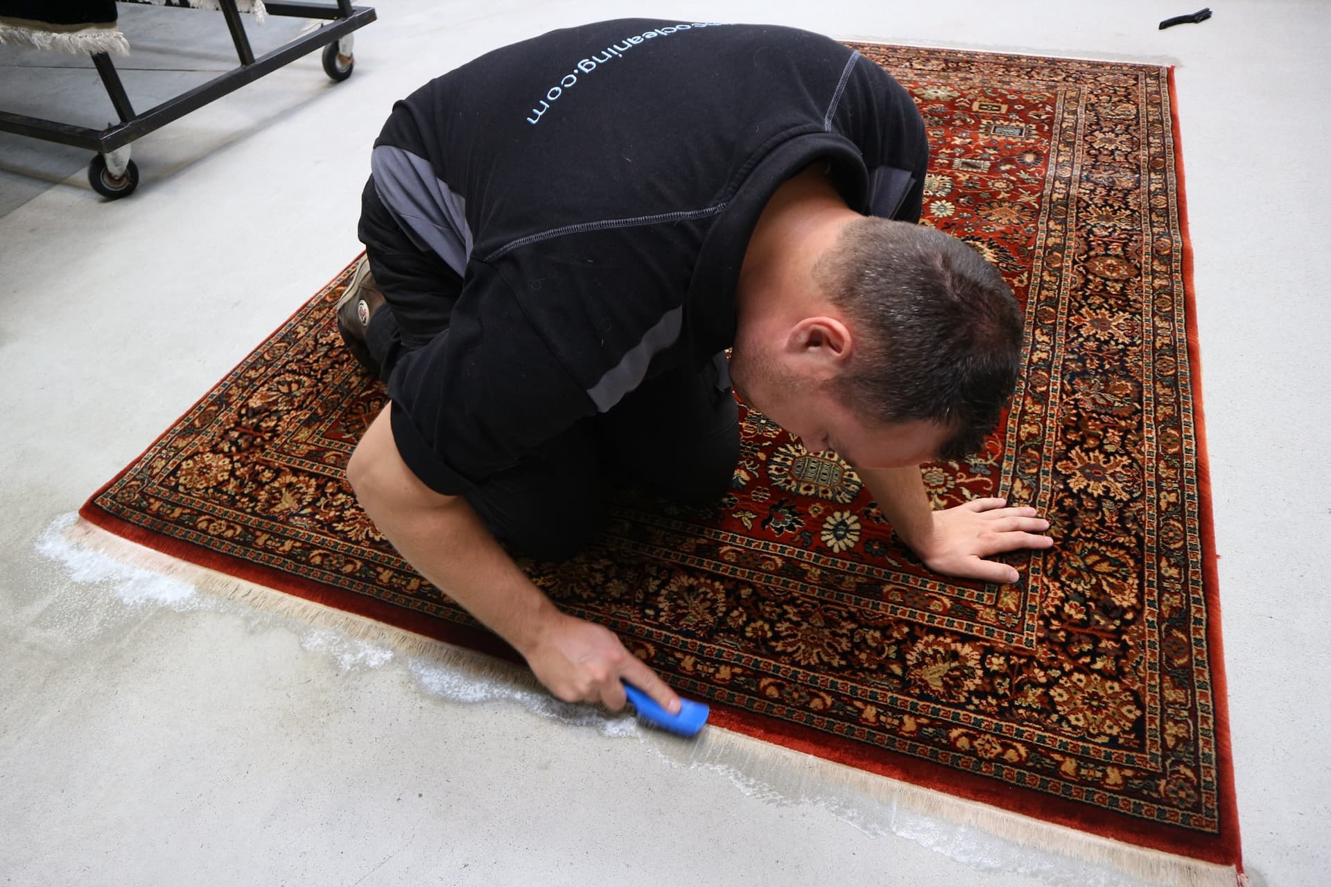 Specialist reinigt knielend de franjes van een antiek vloerkleed met een handborstel